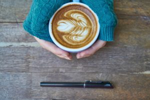 Two hands hold a coffee cup with a pen sitting nearby.